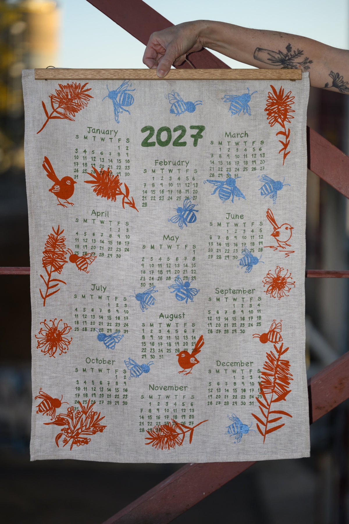 A fabric wall calendar hanging outdoors, showing the year 2027 with monthly grids and printed bird, bee, and floral illustrations in orange, blue, and green. A person's tattooed arm is holding the top edge of the calendar.