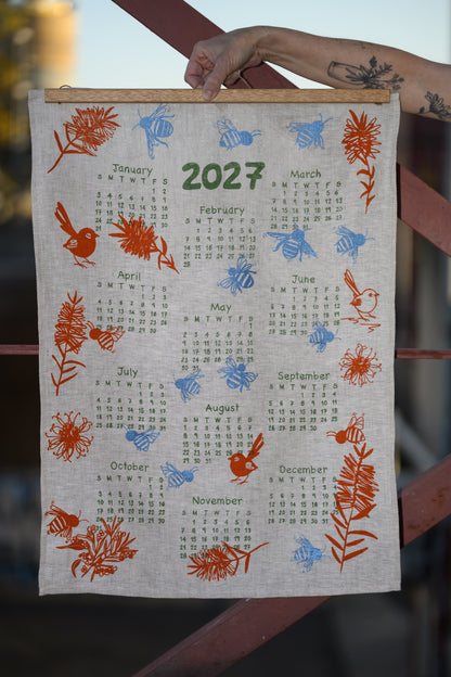 A fabric wall calendar hanging outdoors, showing the year 2027 with monthly grids and printed bird, bee, and floral illustrations in orange, blue, and green. A person's tattooed arm is holding the top edge of the calendar.
