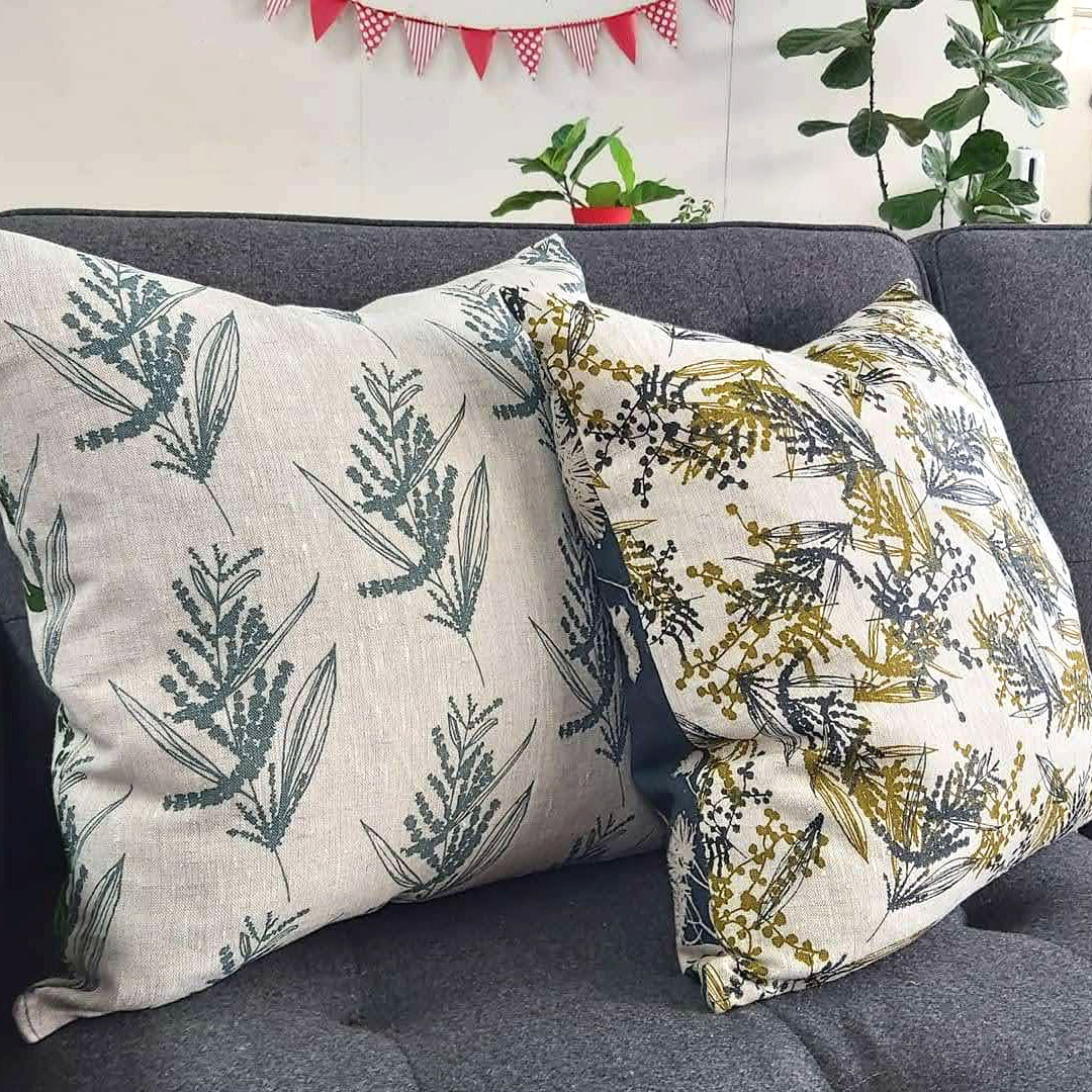 Two linen cushion covers on a couch featuring wattle print in storm and mixed wattle in olive oil and indigo. Designed and printed and made by Femke Textiles.